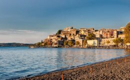 Scenic view of a lakeside village with colorful buildings on a hill, surrounded by calm water and sandy beach, under a clear blue sky.