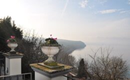Scenic view of a misty lake surrounded by hills, featuring ornamental flower pots on a balcony railing, ideal for nature lovers and travel enthusiasts.