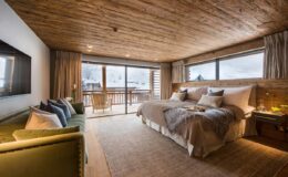 Modern hotel room featuring a king-sized bed, wooden ceiling, and large windows with mountain views. Cozy seating area with green sofa and decorative pillows, complemented by warm lighting and neutral decor. Ideal for a relaxing getaway in a scenic location.