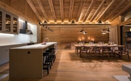 Modern kitchen and dining area featuring wooden beams, a spacious island with bar stools, and a large dining table set for a meal, illuminated by stylish pendant lights, showcasing a warm and inviting atmosphere.