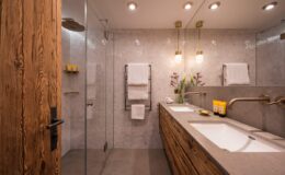 Modern bathroom design featuring a glass shower, dual sinks with a wooden vanity, stylish lighting fixtures, and elegant gray tiles.