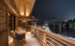Cozy wooden balcony overlooking a snowy landscape at night, with warm lighting and comfortable seating, ideal for relaxation in a mountain setting.