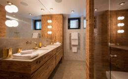 Modern bathroom design featuring wooden cabinetry, stylish lighting fixtures, and a sleek glass shower. The space includes a double sink with ample counter space, neatly arranged towels, and natural light from windows, creating a warm and inviting atmosphere.