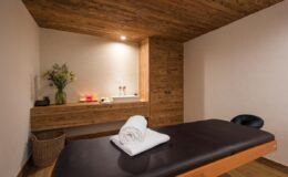Serene spa treatment room featuring a massage table with rolled towels, wooden accents, and soft lighting, ideal for relaxation and wellness services.