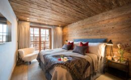 Modern bedroom with wooden ceiling and walls, featuring a large bed with decorative pillows, a cozy armchair, and a side table with flowers, illuminated by natural light from a balcony window.