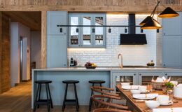 Modern kitchen design featuring light blue cabinetry, a white tiled backsplash, and a large island with seating. The space includes wooden accents, stylish pendant lighting, and a dining area set with tableware, creating a warm and inviting atmosphere.