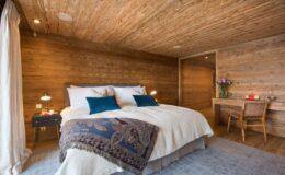 Cozy wooden bedroom featuring a queen-sized bed with decorative pillows, a bedside table with lamps, a rustic wooden desk, and natural light from large windows, creating a warm and inviting atmosphere.