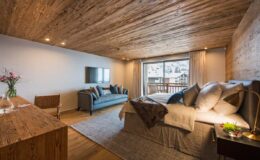 Cozy modern bedroom featuring a wooden ceiling, plush bedding, and a comfortable seating area with a sofa, complemented by natural light from large windows and elegant decor.