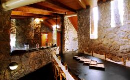 Interior view of a rustic stone dining area featuring a long black table, wooden beams, and natural light streaming through small windows, creating a warm and inviting atmosphere.