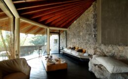 Modern living room with stone walls, wooden beams, and large windows, featuring minimalist furniture and a cozy atmosphere.