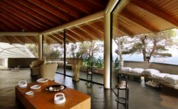 Modern interior of a spacious living room featuring large glass windows, wooden beams, and a minimalist design. The room showcases a wooden coffee table with decorative items, comfortable seating, and a serene view of nature outside, creating a tranquil atmosphere.
