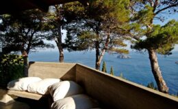 Cozy outdoor seating area with cushions, surrounded by trees, overlooking a serene ocean view on a sunny day. Ideal for relaxation and enjoying nature.