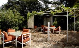 Outdoor dining area featuring a stone patio, white metal furniture with orange cushions, and a pergola adorned with greenery, surrounded by lush trees. Ideal for relaxing and entertaining in a natural setting.