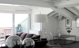 Modern living room with a stylish black sofa, glass coffee table, and large floor lamp, featuring exposed wooden beams and ample natural light from large windows. Decorative elements include silver spheres and a vase with flowers.