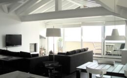 Modern living room with high ceilings, large windows, and a cozy seating area featuring a black sofa, coffee table, and flat-screen TV, creating a bright and airy atmosphere.