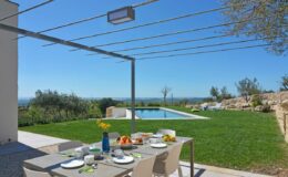 Outdoor dining area with a table set for breakfast, featuring plates, fruits, and a blue vase, overlooking a serene pool and green lawn under a clear blue sky.