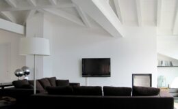 Modern living room featuring a dark sectional sofa, a sleek floor lamp, and a flat-screen TV mounted on a white wall. The space boasts high, exposed beam ceilings and minimalistic decor, creating a bright and airy atmosphere.