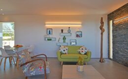 Modern living room featuring a green sofa with decorative pillows, a wooden sculpture, and a dining area with stylish chairs. The space is illuminated by soft lighting and decorated with wall shelves displaying plants and art.