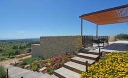 Modern outdoor terrace with stone wall, steps, and vibrant flowers, overlooking a scenic landscape under a clear blue sky.