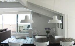 Modern dining area with a wooden table, white chairs, and stylish pendant lights, featuring a minimalist design and large windows that provide natural light and a view of an outdoor balcony.