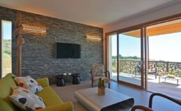 Modern living room featuring a green sofa with decorative pillows, a stone accent wall, a mounted television, and large glass doors leading to a balcony with scenic views.