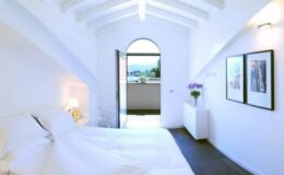 Modern bedroom with white walls, a sloped ceiling, and a large window opening to a balcony. The room features a double bed with white linens, a bedside lamp, and two framed pictures on the wall, creating a bright and airy ambiance.