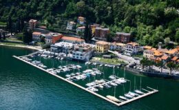 Aerial view of a picturesque lakeside marina with yachts docked, surrounded by colorful buildings and lush greenery. Ideal for travel and boating enthusiasts.