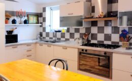 Modern kitchen with a wooden dining table, featuring a checkerboard backsplash, stainless steel appliances, and ample storage space. Brightly lit with natural light from a window, showcasing a stylish and functional design.