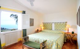 Cozy bedroom with a green and yellow patterned bedspread, featuring a double bed, bedside lamps, and a scenic ocean view through an open window. Bright and airy atmosphere with natural light, wooden flooring, and stylish decor. Perfect for a relaxing getaway.