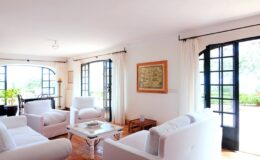 Bright and airy living room featuring white sofas, large windows with black frames, and a wooden coffee table. Natural light fills the space, highlighting the minimalist decor and indoor plants.