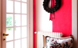 Cozy entryway featuring a vibrant red wall, a decorative wreath with a ribbon, a radiator, and a plush floral-patterned cushion on a wooden chair, creating a welcoming atmosphere.