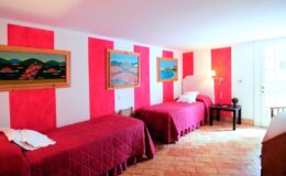 Bright and inviting bedroom featuring two twin beds with red bedspreads, colorful landscape paintings on the wall, and natural light streaming in from a window.