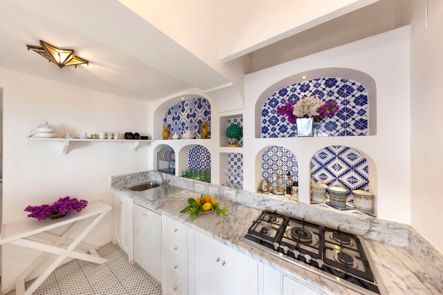 Bright and modern kitchen featuring white cabinetry, a marble countertop, and decorative blue and white tile accents. The space includes a sink, stove, and shelves displaying colorful flowers and kitchenware, creating a vibrant and inviting atmosphere. | Only on SopranoVillas