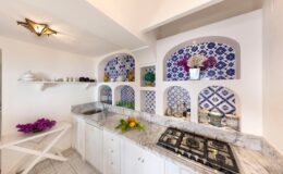 Bright and modern kitchen featuring white cabinetry, a marble countertop, and decorative blue and white tile accents. The space includes a sink, stove, and shelves displaying colorful flowers and kitchenware, creating a vibrant and inviting atmosphere.