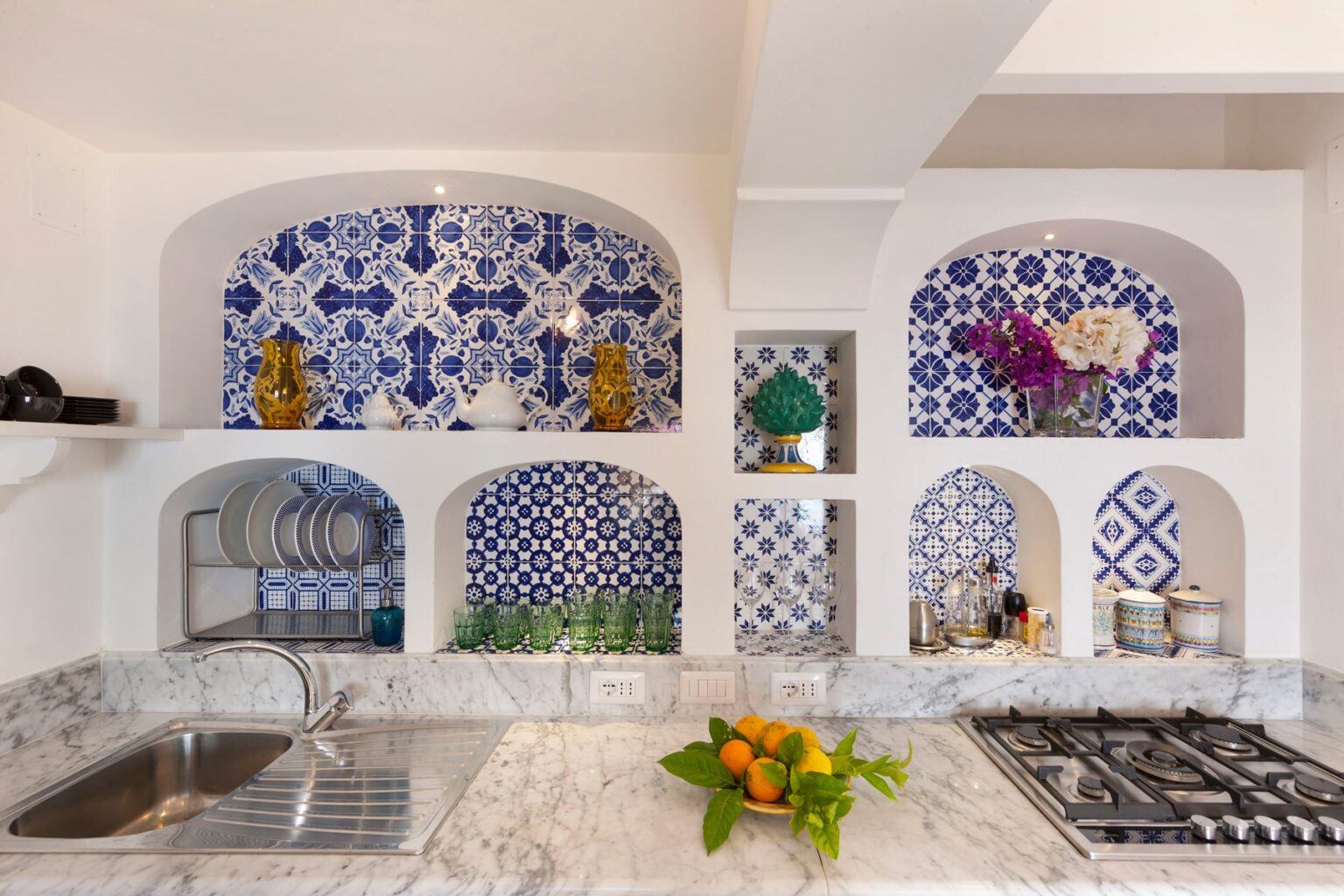 Bright and modern kitchen featuring blue and white patterned tile accents, decorative niches with plants and vases, marble countertops, and a stainless steel gas stove. | Only on SopranoVillas