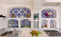 Bright and modern kitchen featuring blue and white patterned tile accents, decorative niches with plants and vases, marble countertops, and a stainless steel gas stove.