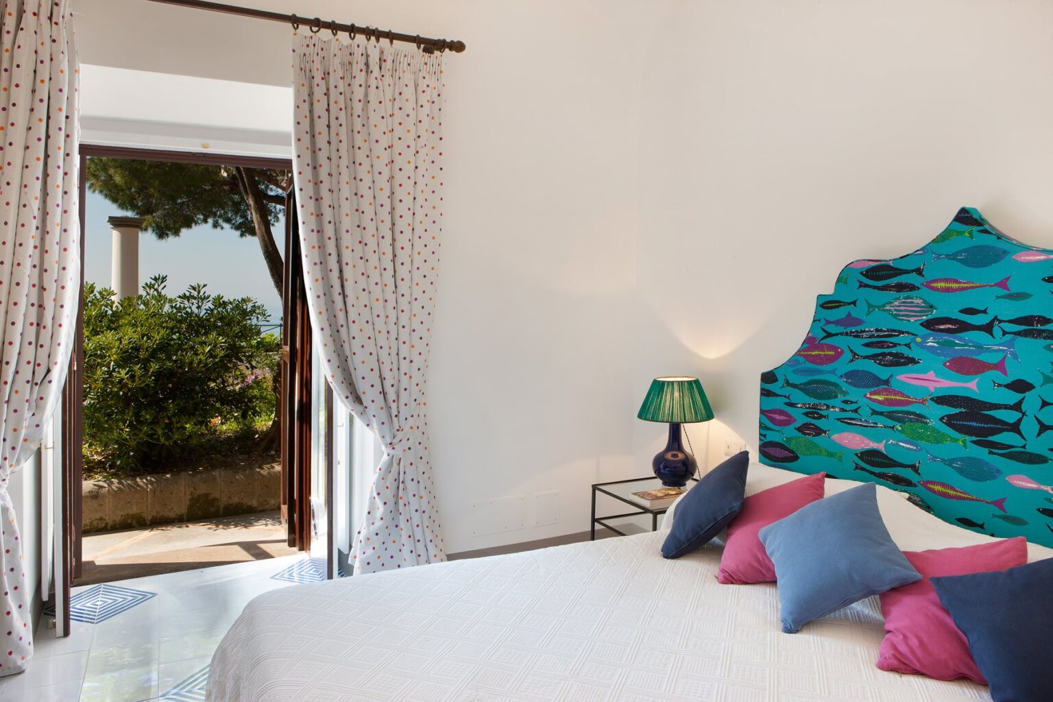 Bright and stylish bedroom with a colorful headboard, decorative pillows, a bedside lamp, and a view of a garden through open doors. Ideal for showcasing interior design and cozy living spaces. | Only on SopranoVillas