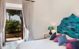 Bright and stylish bedroom with a colorful headboard, decorative pillows, a bedside lamp, and a view of a garden through open doors. Ideal for showcasing interior design and cozy living spaces.