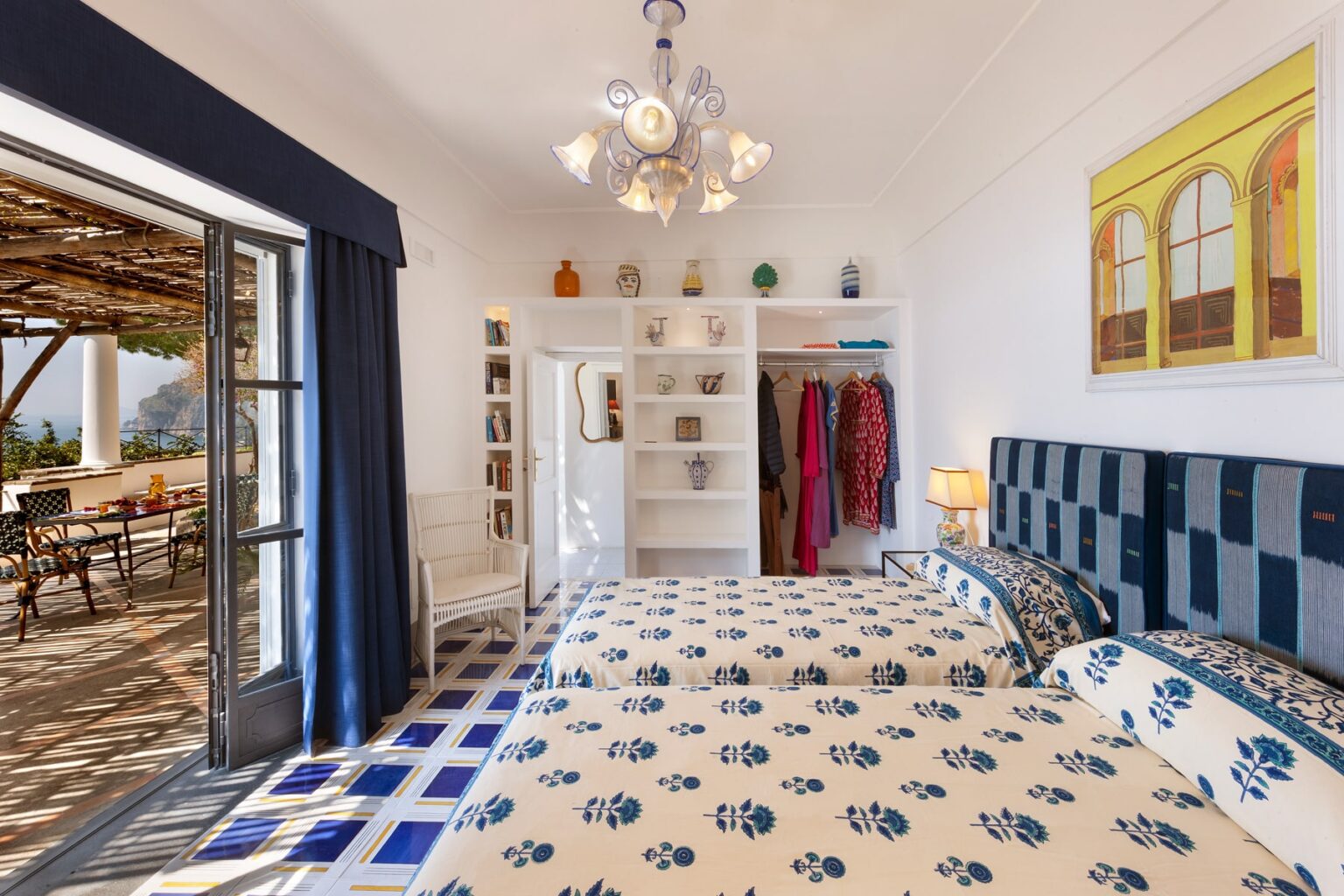 Cozy bedroom featuring two twin beds with floral bedding, a stylish chandelier, and a bright window leading to an outdoor patio. The room includes a white shelving unit and vibrant wall art, creating a welcoming atmosphere. | Only on SopranoVillas