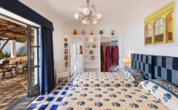 Cozy bedroom featuring two twin beds with floral bedding, a stylish chandelier, and a bright window leading to an outdoor patio. The room includes a white shelving unit and vibrant wall art, creating a welcoming atmosphere.