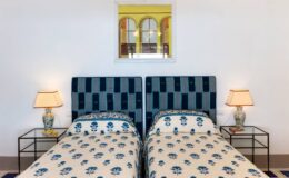 Cozy bedroom featuring two twin beds with patterned bedding, stylish bedside lamps, and a decorative mirror above. Bright and inviting atmosphere with a touch of elegance.