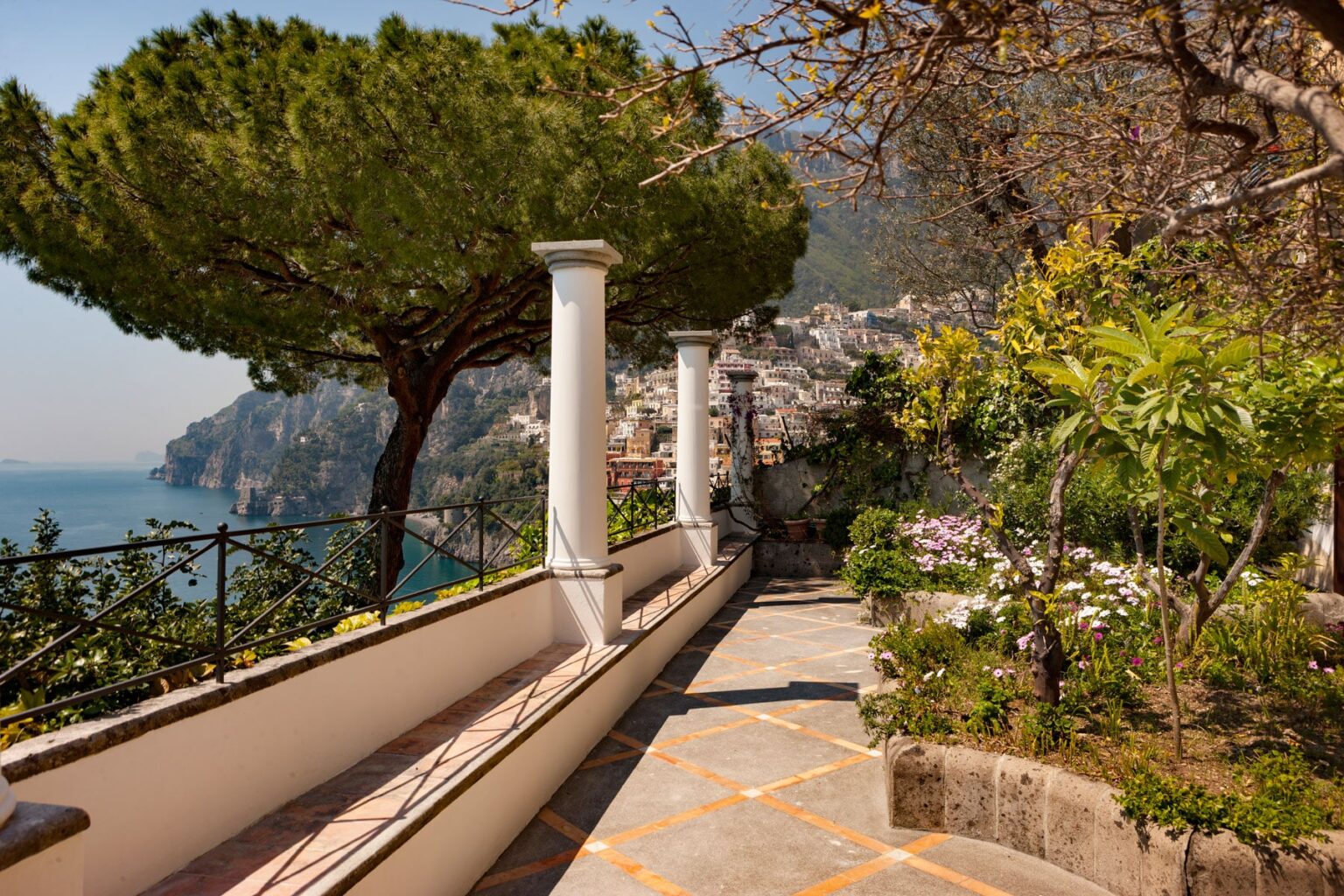Scenic pathway lined with lush greenery and tall trees, overlooking the coastline of a picturesque village, with mountains in the background and a clear blue sky. | Only on SopranoVillas