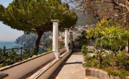 Scenic pathway lined with lush greenery and tall trees, overlooking the coastline of a picturesque village, with mountains in the background and a clear blue sky.