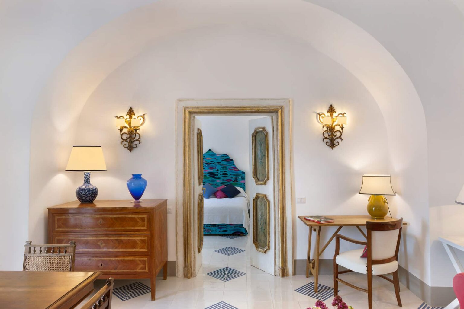 Elegant interior of a modern living space featuring arched architecture, decorative wall sconces, a wooden dresser, and a stylish desk. A glimpse of a bedroom with turquoise decor is visible through the doorway. | Only on SopranoVillas