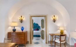 Elegant interior of a modern living space featuring arched architecture, decorative wall sconces, a wooden dresser, and a stylish desk. A glimpse of a bedroom with turquoise decor is visible through the doorway.