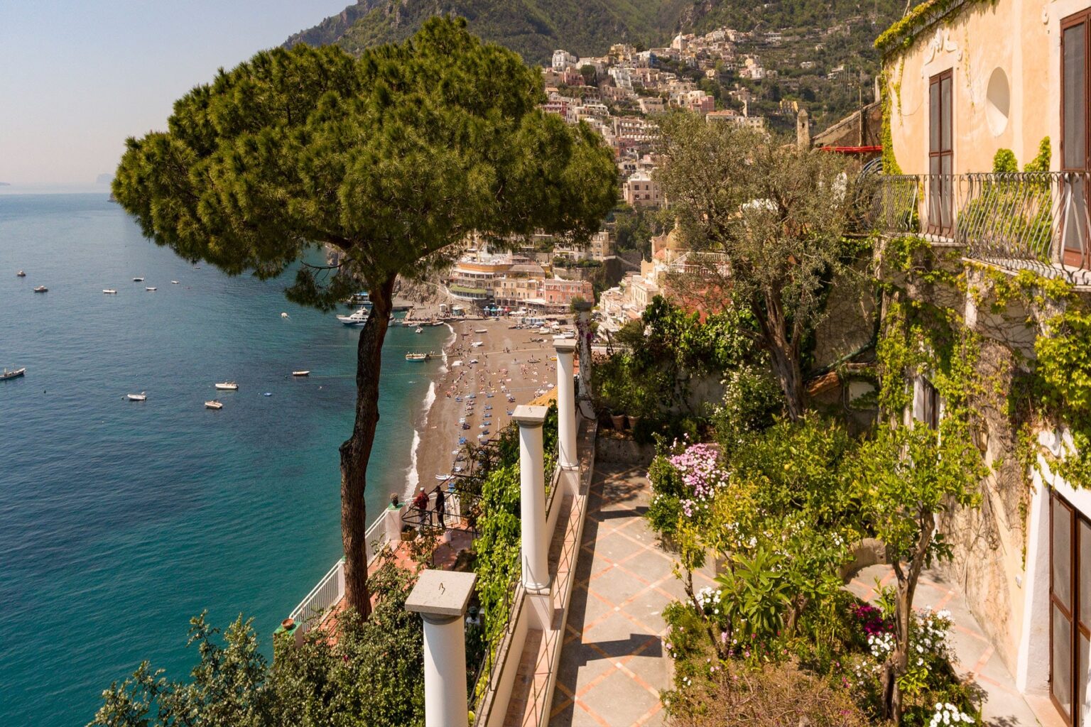 Scenic view of a coastal pathway lined with lush greenery, overlooking a beach and clear blue waters, with charming buildings and hills in the background. | Only on SopranoVillas
