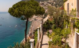 Scenic view of a coastal pathway lined with lush greenery, overlooking a beach and clear blue waters, with charming buildings and hills in the background.