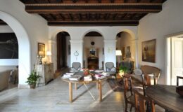 Spacious interior of a stylish living room featuring arched doorways, wooden beams, and a round table with books. The room is well-lit with modern lamps and includes comfortable seating, plants, and art on the walls, creating an inviting atmosphere.