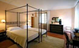 Cozy bedroom featuring a four-poster bed with two twin mattresses, soft lighting, and a stylish dresser. The room is adorned with a neutral color palette and includes a small seating area, creating a warm and inviting atmosphere. Perfect for relaxation and comfort.