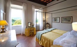 Cozy hotel room featuring two twin beds with yellow bedspreads, large windows with sheer curtains, a vintage dresser, and warm lighting, creating a serene and inviting atmosphere.